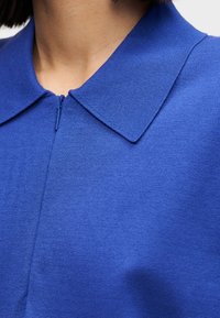 Φωτεινό μπλε πλεκτό ένδυμα με κλείσιμο φερμουάρ και μυτερό γιακά. Το ύφασμα φαίνεται λείο και ελαφρώς υφασμένο, αναδεικνύοντας τον σχεδιασμό του.