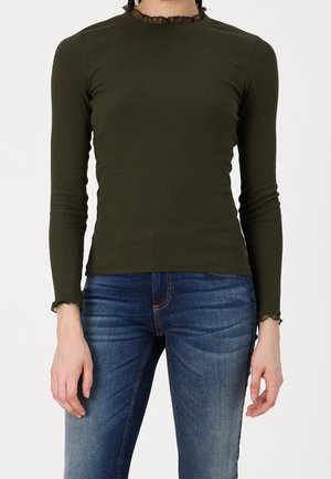 Person wearing a fitted olive green long-sleeve top with ruffled collar and cuffs paired with blue faded jeans, standing against a white background.