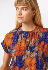 Kortærmet bluse i livlig orange og blå blomstermønster. Har rund halsudskæring, knapper foran og tekstureret stof med plisseringer.