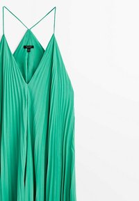 Robe verte plissée sans manches avec de fines bretelles spaghetti et un décolleté en V, suspendue sur un fond blanc.