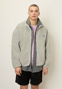 Hellgraue, flauschige Fleecejacke mit hohem Kragen, Reißverschluss vorne und sichtbarem Markenpatch; getragen über einem rosa T-Shirt und dunklen Shorts.