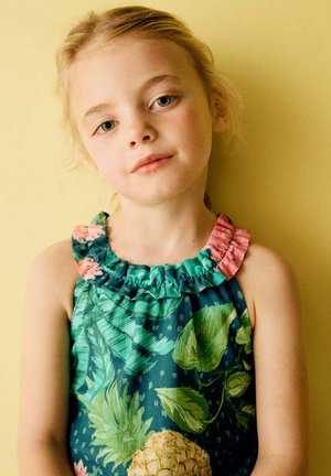 Jeune fille aux cheveux blonds portant une robe sans manches imprimée tropicale verte, debout sur un fond jaune.