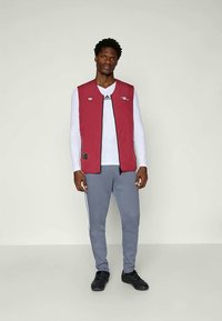 Κερασιές χωρίς μανίκια γιλέκο με μαύρο φερμουάρ και λογότυπα, τοποθετημένο πάνω από ένα λευκό πουκάμισο με μακριά μανίκια και γκρι παντελόνι φόρμας με λευκές ρίγες.