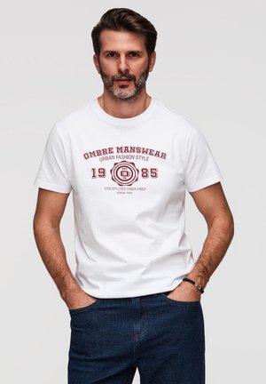 Homme barbu portant un t-shirt blanc avec un motif graphique et un jean bleu, les mains dans les poches, debout devant un fond clair uni.