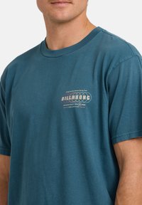 Turkusowy bawełniany t-shirt z okrągłym dekoltem, krótkimi rękawami i graficznym nadrukiem logo na lewej piersi. Miękka tekstura, luźny krój.