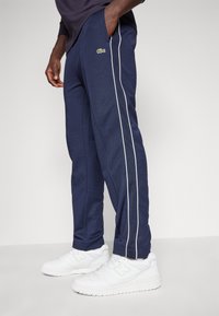 Marineblå trening bukser med hvite side striper, med en grønn Lacoste-logo. Båret med hvite joggesko. Glatt stoff og avslappet passform.