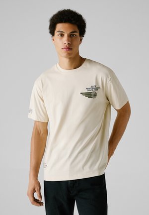 Creme bomulds T-shirt med et grønt grafisk design og teksten "ELITE PERFORMANCE SINCE 2004" samt vingedesign på venstre bryst. Korte ærmer.