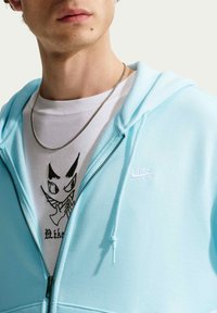 Νεαρός με ασημένιο κολιέ αλυσίδα, λευκό T-shirt με μαύρο καρτούν πρόσωπο και ανοιχτό μπλε φούτερ Nike SB με φερμουάρ.