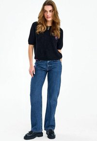 Žena s dugom kosom u crnoj majici kratkih rukava, plavim trapericama ravnih nogavica, crnim debelim cipelama i ogrlicom, na bijeloj pozadini.