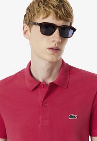 Punainen polo-paitaja, jossa on teksturoitu kangas, kolmella napilla varustettu kaulus ja musta brodeerattu logo rinnassa. Mallilla on päällään ruskeat aurinkolasit.