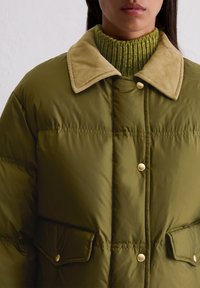 Olivgrüne Steppjacke mit einem samtigen, hellbraunen Kragen, Druckknöpfen und seitlichen Taschen, die eine glatte, gepolsterte Textur aufweist.