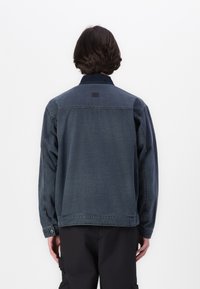 Denim jacket in dark blue with a button-down collar, relaxed fit, and visible stitching details. Features a small brand tag on the back.