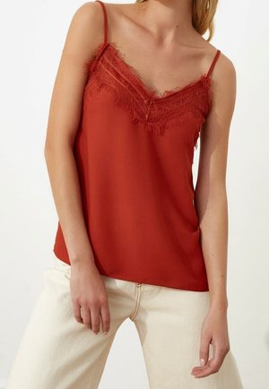 Kvinde iført en ærmeløs rust-rød camisole med blondekant og spaghetti stropper, kombineret med lyse beige bukser.