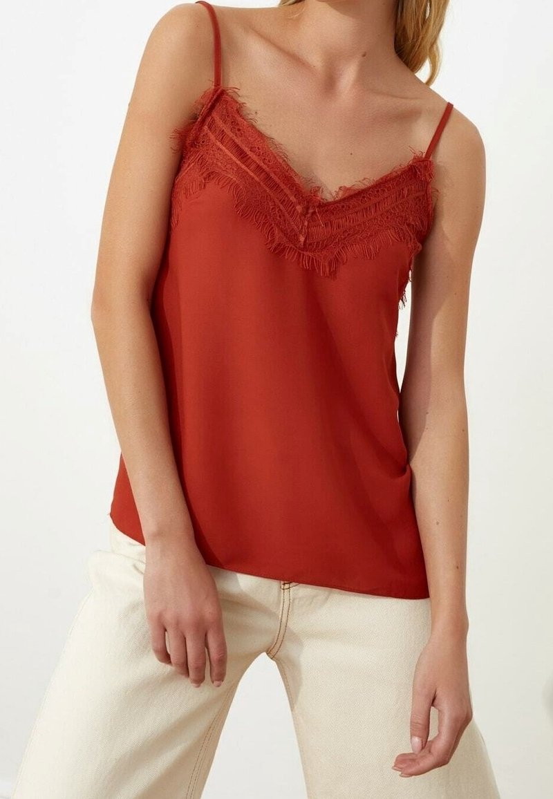 Femme portant un caraco rouge rouille sans manches avec bordure en dentelle et bretelles spaghetti, associé à un pantalon beige clair.