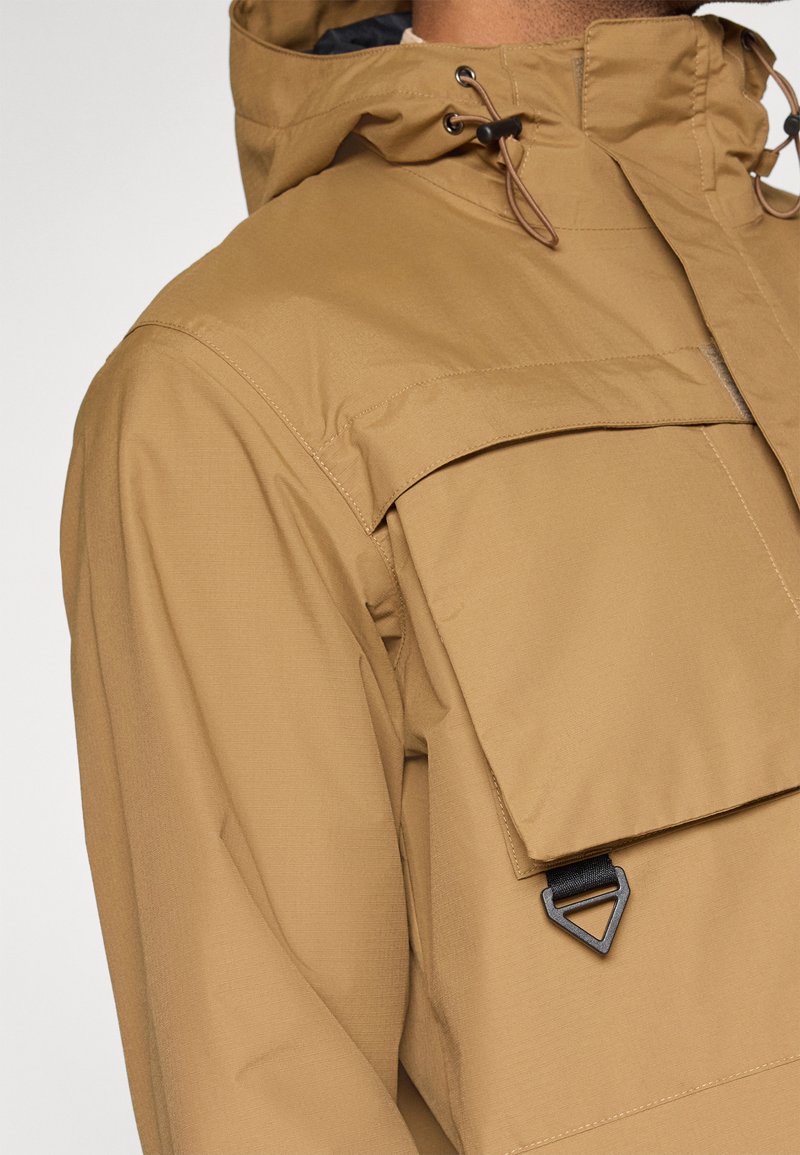 Chaqueta impermeable color beige con capucha, gran bolsillo frontal y acento de hardware negro triangular. Acabado de tela suave y texturizada.