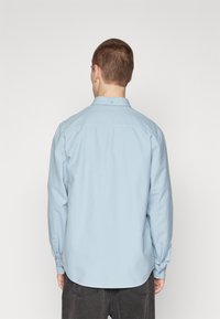 Chemise bleu clair à manches longues avec col, fermeture à boutons et détail de pli à l'arrière. Le tissu semble lisse et léger.
