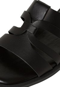 Sandale en cuir noir avec plusieurs brides et un accent noeud. Elle présente une texture lisse et un design ouvert pour la respirabilité. Semelle plate.