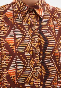 Camicia marrone a fantasia con motivi geometrici arancioni, gialli e neri, dotata di colletto a punta e chiusura con bottoni in legno.
