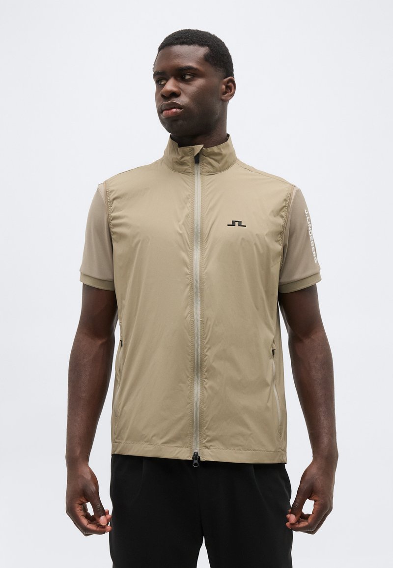 Άνδρας με μπεζ αμάνικο γιλέκο με φερμουάρ πάνω από κοντομάνικο πουκάμισο, στέκεται μπροστά σε απλό λευκό φόντο, κοιτώντας προς τα αριστερά.