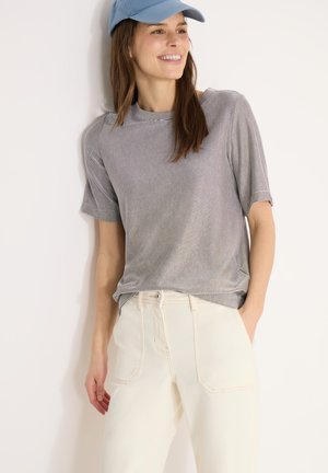 Ung kvinde smilende, iført lyseblå kasket, gråt stribet kortærmet top og cremefarvede højtaljede bukser, stående op ad en hvid væg.