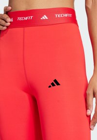 Røde Techfit leggings med et glat, strækbart stof, en bred talje med hvid branding og et sort logo nær anklen.