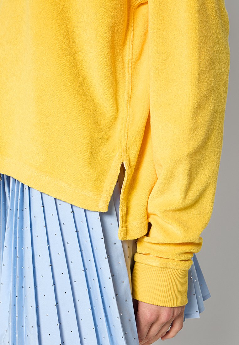 Sweatshirt in pile giallo con vestibilità rilassata, fessure laterali e polsini a costine, abbinato a una gonna a pieghe azzurro chiaro con piccoli dettagli in argento.