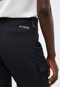 Primer plano de una persona con pantalones negros Columbia Titanium, donde se ve el bolsillo trasero y el lazo para el cinturón, con la mano apoyada cerca del muslo.