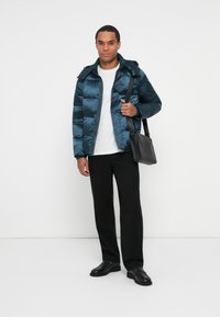 Hombre vestido con chaqueta acolchada azul, camisa blanca, pantalones negros, mocasines negros y llevando un bolso de hombro negro, contra un fondo blanco.