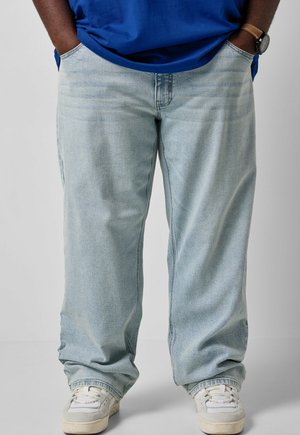 Persona che indossa jeans azzurri a gamba larga, sneaker bianche, una camicia blu e un orologio da polso, con le mani in tasca.