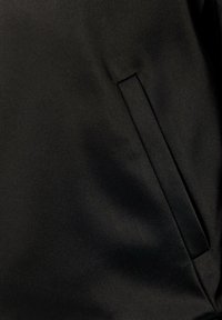 Zwarte stof met een gladde textuur, voorzien van een enkele smalle zijzak die in het ontwerp is geïntegreerd. Minimalistische en moderne uitstraling.