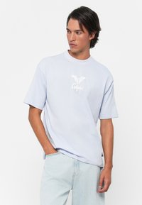 Ung mann iført en lyseblå t-skjorte med en hvit sommerfugl-logo og lyseblå jeans, som ser til siden med den ene hånden i lommen.