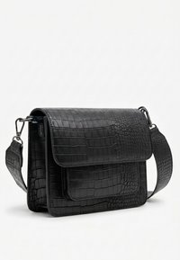 Zwarte faux krokodillenleren crossbodytas met klepsluiting en verstelbare brede schouderband op een witte achtergrond.