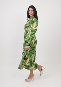 Abito floreale verde con ampi motivi a foglia, maniche lunghe, fascia da annodare in vita e gonna fluida. Indossato con sandali con tacco beige.