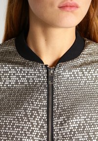 Primo piano di una persona che indossa una giacca a motivi argento e nera con colletto nero e una cerniera centrale argentata.