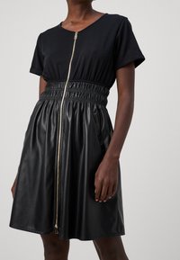 Schwarzes Kleid mit einem kurzärmeligen Baumwolloberteil und einem gerafften Rock aus Kunstleder. Zentraler Reißverschluss und seitliche Taschen sind vorhanden.