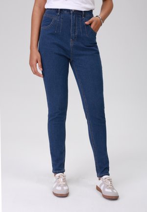 High-waisted, slim-fit jeans in dark blue denim with visible stitching and front pockets, paired with light gray sneakers.