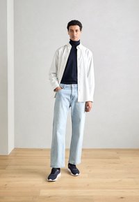 Chemise blanche à boutons portée par-dessus un turtleneck bleu marine, jean droit bleu clair et baskets bleu marine avec semelles blanches, debout sur un sol en bois.