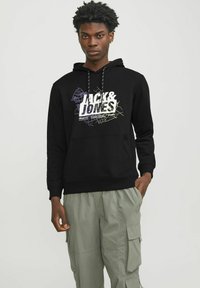 Sudadera negra con diseño gráfico blanco y colorido 'JACK & JONES', capucha con cordón, bolsillo frontal, llevada con pantalones cargo de color verde claro.
