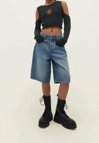 Person wearing a black ripped cropped sweater, loose knee-length denim shorts, and black chunky lace-up boots on a light background.