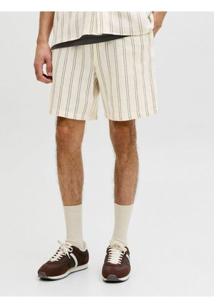 Férfi krémszínű, vékony fekete függőleges csíkos rövidnadrágban, fehér zokniban, barna tornacipőben, egyik kezét zsebében tartva áll.