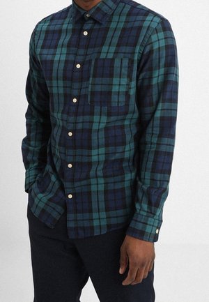 Mann, der ein grün-kariertes Hemd mit dunkelblauen Karos, langen Ärmeln und schwarze Hose trägt, steht mit einer Hand in der Tasche.