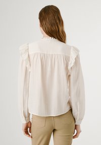 Helle Cremebluse mit Rüschendetails an den Schultern, gerafftem Rücken und langen Ärmeln, aus einem weichen, leichten Stoff hergestellt.