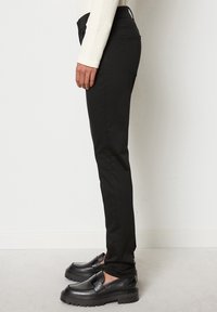 Pantaloni neri slim-fit con una texture liscia, caratterizzati da una vita media, tasche posteriori e abbinati a mocassini neri chunky.
