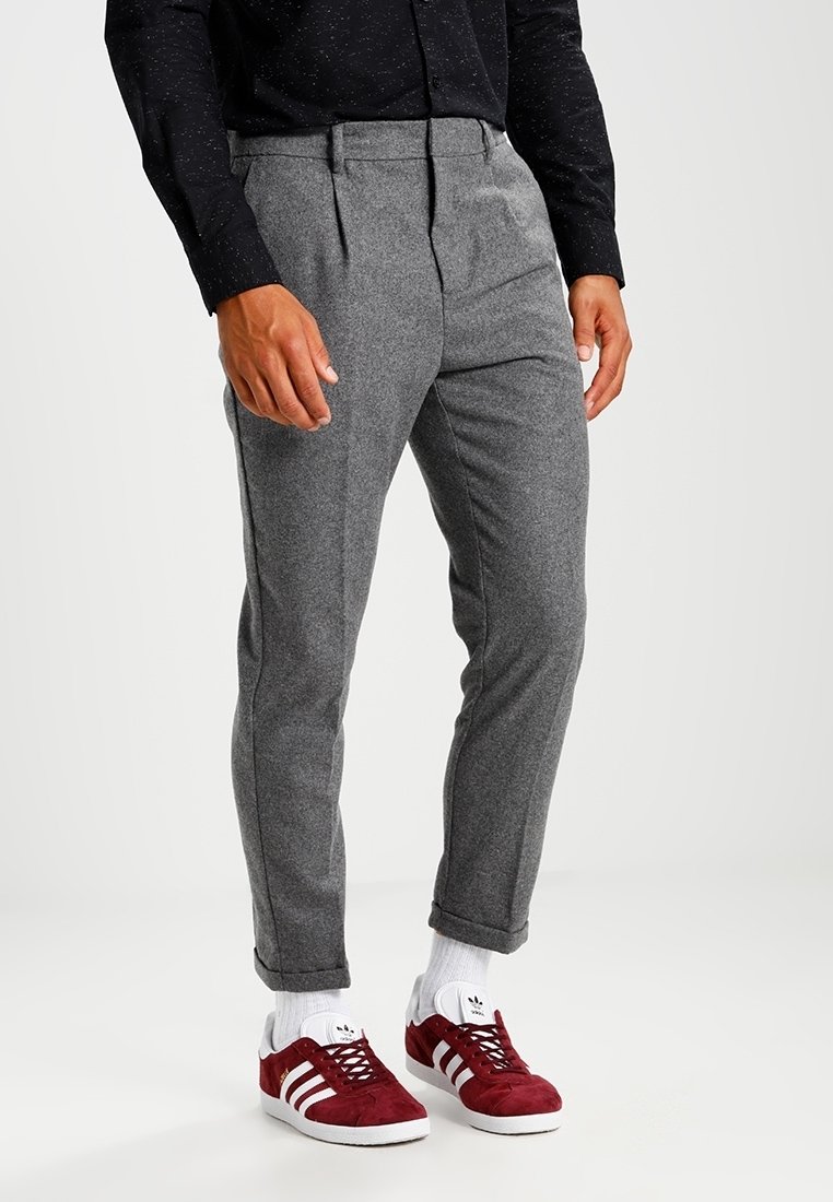 Homme portant un pantalon gris sur mesure retourné aux chevilles, des chaussettes blanches et des baskets rouges Adidas avec des bandes blanches.