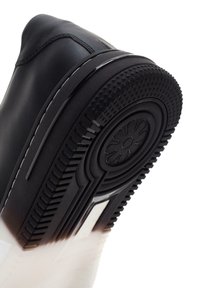 Chaussure de sport noire avec une tige en cuir lisse, semelle en caoutchouc dotée d'un motif de grip texturé et d'un motif floral au centre.