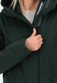 Tmavě zelená bunda na zip s kapucí, s viditelnou značkovou etiketou a pruhovanou vnitřní košilí. Měkká textura a žádné viditelné vzory.