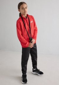 Crvena sportska jakna sa zatvaračem i visokim ovratnikom, crne trenirke i crne atletske cipele. Odjeća sadrži Puma logo.