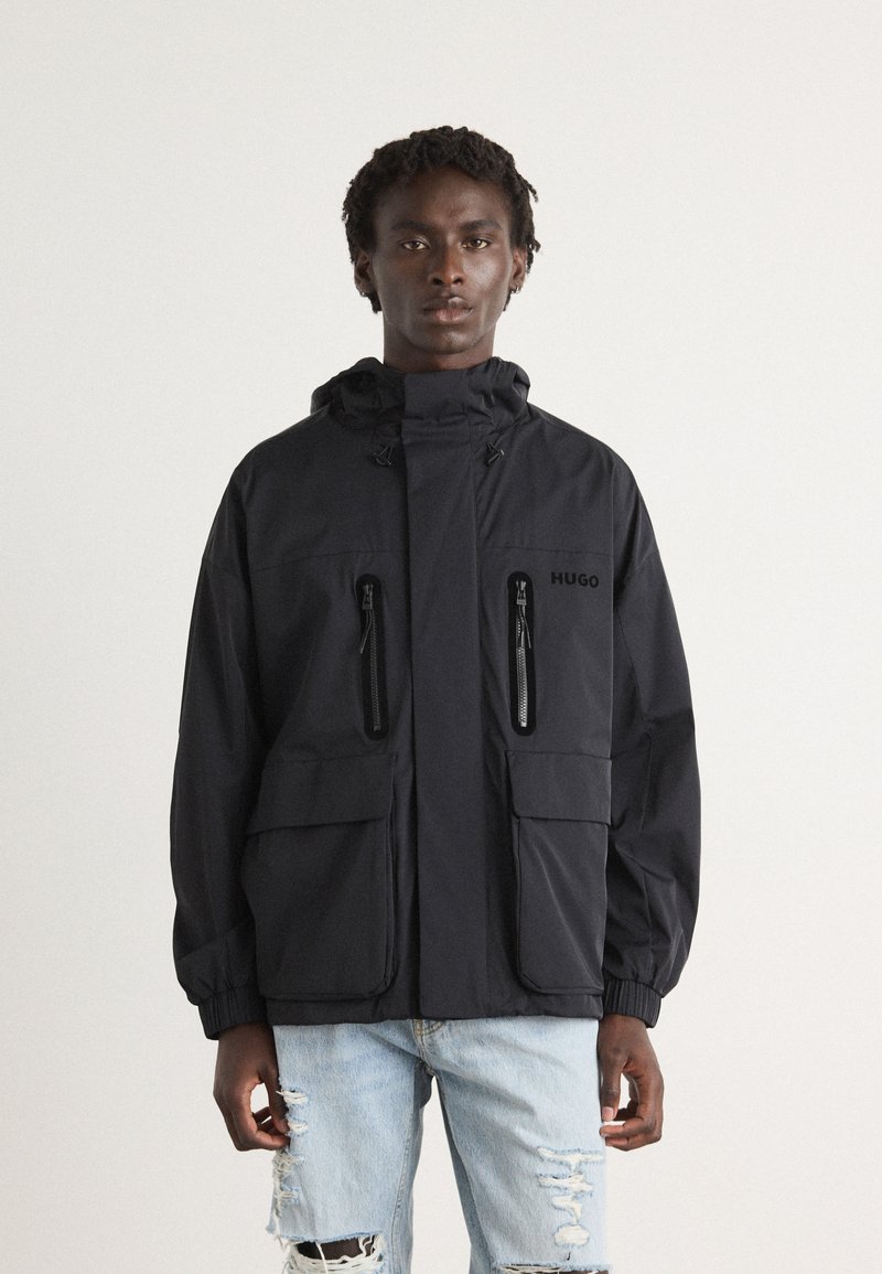 Black hooded jacket with two zippered pockets, elastic cuffs, and a logo on the left chest. Worn over distressed light blue jeans.