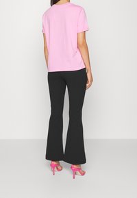 Kvinna i en rosa kortärmad tröja, svarta utsvängda byxor och rosa högklackade sandaler som står mot en helt vit bakgrund.