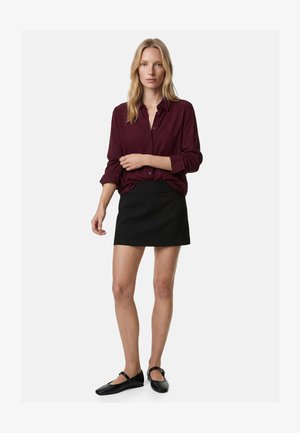 Kobieta stoi, ubrana w bordową koszulę z długim rękawem, czarną mini spódniczkę oraz czarne baleriny Mary Jane na jednolitym jasnym tle.